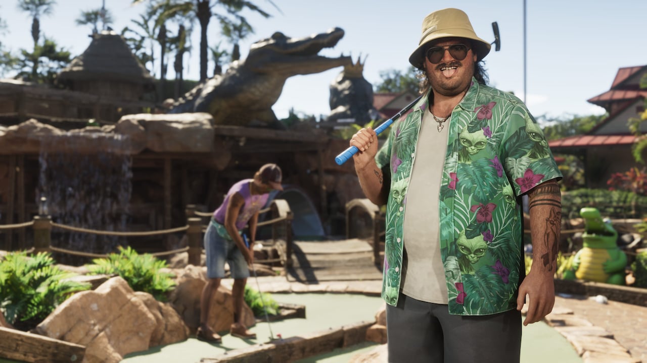 A grinning man in a green shirt printed with alien faces and pink hibiscus flowers holds a putter over his shoulder at a Leonida mini-golf course in official GTA VI artwork, with a giant crocodile statue and a waterfall behind him and a second player putting on the green