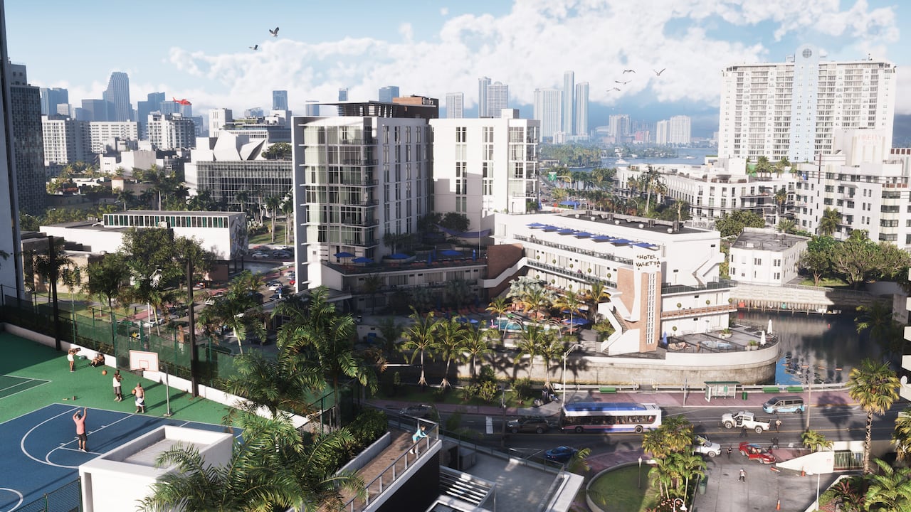Daytime aerial of a Vice City beachfront block in official GTA VI artwork, showing a hotel pool deck lined with rows of blue umbrellas and loungers, palm trees, a public basketball court with players, a bus on the boulevard and the downtown skyline behind